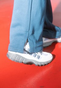 Τελείες αθλητικές παντελόνες σε τυρκουάζ απόχρωση με πλαϊνές σχισμές, σε συνδυασμό με άσπρα αθλητικά παπούτσια με μπλε και γκρι λεπτομέρειες και υφασμένη μαύρη σόλα.