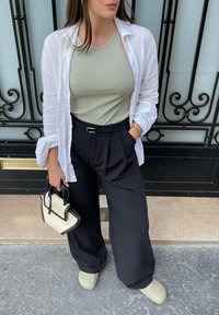 Lichtgroene geribde tanktop in combinatie met een witte linnen overhemd, zwarte wijde broek en beige clogs. Houdt een kleine zwart-wit handtas vast.