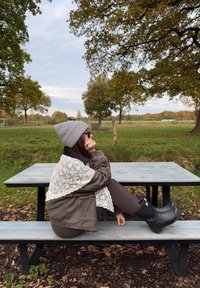 Brown leather jacket with white lace trim, gray knitted beanie, black rubber boots. Sitting on a wooden picnic table in a natural setting.