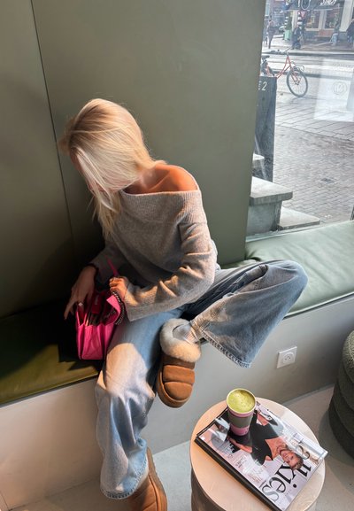 Jersey gris de hombros descubiertos, vaqueros de pierna ancha azul claro, zapatillas marrones de piel de oveja. Sosteniendo un bolso rosa brillante. Bebida verde sobre una mesa con una revista.