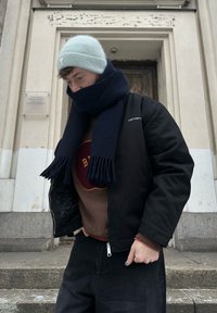 Černá bombardérka se zipovým zapínáním, hnědý svetr s kruhovým logem, námořnický šál s třásněmi a světle modrá pletená čepice.