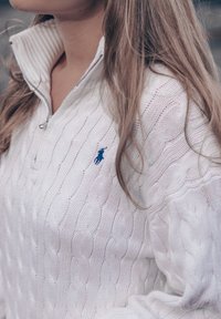 Hvit kabelstrikket genser med en halvlengdes glidelåskrage, med en liten blå logo på brystet. Myk tekstur med ribbestrikket kragedetalj.