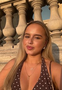 Ruskea pilkullinen solmittava toppi, herkkä kultainen kaulakoru, jossa on rusetti ja riipus, pitkät vaaleat hiukset, pehmeä auringonvalo, teksturoitu kivikaide taustalla.