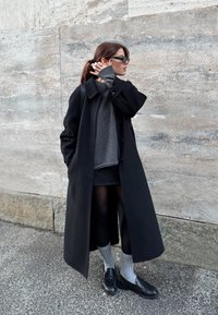 Γυναίκα που φοράει μακρύ μαύρο παλτό, γκρι πουλόβερ, μαύρη φούστα, γκρι κάλτσες και μαύρα παπούτσια, στέκεται δίπλα σε πέτρινο τοίχο, διορθώνοντας τα μαλλιά της με γυαλιά ηλίου.