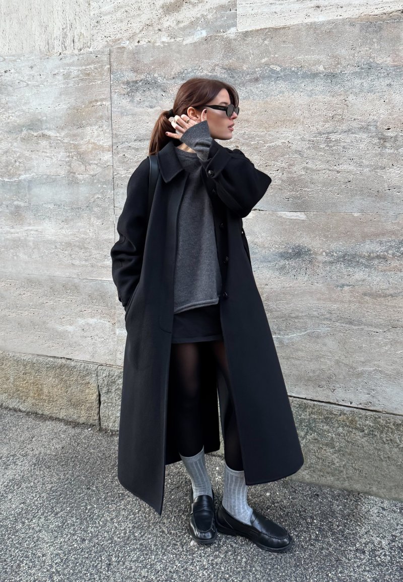 Donna con indosso un lungo cappotto nero, maglione grigio, gonna nera, calzini grigi e scarpe nere, in piedi contro un muro di pietra, si sistema i capelli con gli occhiali da sole indossati.