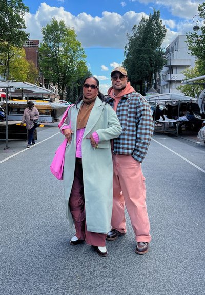 Dos personas están de pie en una calle. Una viste un abrigo verde menta, una camiseta rosa, pantalones marrones y sostiene un bolso rosa. La otra lleva una chaqueta a cuadros y pantalones rosas.
