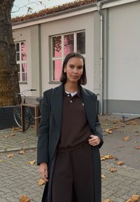 Fekete, túlságosan nagy blézer barna galléros póló és hozzáillő nadrág felett. Arany karika fülbevalók. A háttérben fa és kerékpár látható. Őszi levelek a járdán.