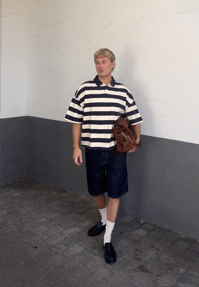 Joven caminando en el interior con una camisa a rayas negras y blancas, pantalones cortos oscuros, calcetines blancos, zapatos negros y llevando una bolsa marrón.