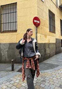 Černá kožená bunda s ovčí vlnou, šedá mikina, kostkovaná košile svázaná kolem pasu a šedé džíny, na pozadí kamenn�é ulice.