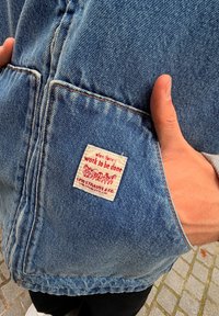 Denimjakke med lysblåt stof, synlige syninger og et rødt og hvidt vævet mærke på lommen, der siger "når der er arbejde, der skal gøres."