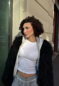 Svart lodden kåpe over en grå hettegenser og hvit tettsittende topp, kombinert med grå joggebukser. Krøllete hår, naturlig sminke, mykt sollys.