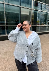 Grijze wollen cardigan over een wit shirt, zwarte broek met witte stippen, doorzichtige zonnebril en een kleine zilveren schoudertas.