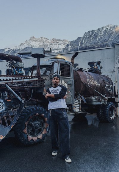 Hombre con ropa casual de pie con los brazos cruzados frente a un vehículo todoterreno grande y muy modificado con un fondo de montañas.