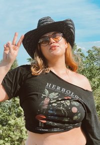 Μαύρη κοντή μπλούζα με γραφικό "Ερμπολογία" που απεικονίζει φυτά, φορεμένη στον ένα ώμο. Αξεσουάρ περιλαμβάνουν μια μαύρη καπέλο από ψάθα και γυαλιά ηλίου.