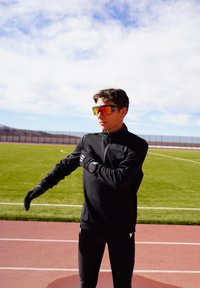 Athlète en tenue de sport noire et lunettes de soleil réfléchissantes s'étire le bras sur une piste de course extérieure sous un ciel partiellement nuageux.