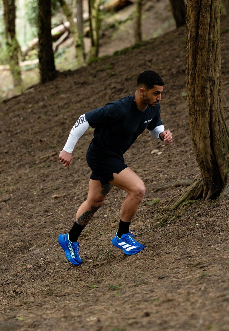 Man draagt blauwe hardloopschoenen en zwarte sportkleding en rent heuvelop op een bospad nabij bomen met een geconcentreerde blik.