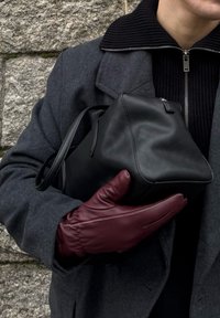 Zwarte leren tas met een gestructureerde vorm, vastgehouden door een hand in een bordeauxrode leren handschoen. Donkergrijze jas en geribde zwarte kraag zichtbaar.