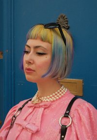 Κοντό ροζ φόρεμα με πούλιες και φουσκωτά μανίκια, μαύρη θηλιά, κολιέ με πέρλες, πολύχρωμο καρέ κούρεμα και μαύρα γυαλιά ηλίου resting στο κεφάλι.