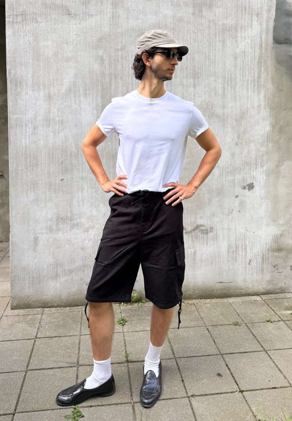 Jeune homme portant un t-shirt blanc, un short noir, des chaussettes blanches, des chaussures noires, une casquette et des lunettes de soleil, debout les mains sur les hanches devant un mur gris.