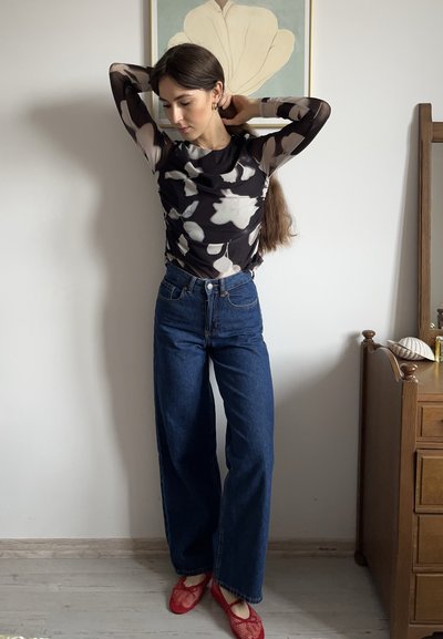 Top negro de manga larga con un patrón floral blanco, combinado con jeans azul de cintura alta y zapatos planos de malla rojos. Pose de pie en un fondo simple.