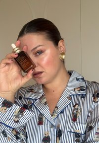 Transparent glassflaske med Estee Lauder-serum med gulldetaljer, holdt mot en blå og hvit stripete pysjamastrøye med små bjørnmønstre.