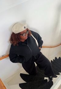 Giacca nera con zip e pantaloni della tuta, berretto bianco con logo. In piedi su scale a spirale scure contro un muro bianco.