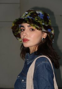 Φανταχτερό καπέλο μπούκλας σε πράσινους, μπλε και καφέ τόνους. Τζην πουκάμισο με κουμπιά σnap. Ορατό μπεζ λουράκι ώμου. Φυσικό μακιγιάζ και σκουλαρίκια.