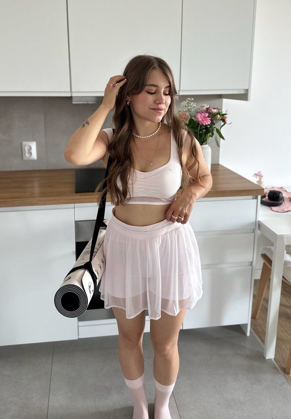 Lys rosa sports-bh med mesh-detaljer, lagd gjennomsiktig skjørt og knelengde rosa sokker. Bærer en rullet yogamatte med svart stropp.