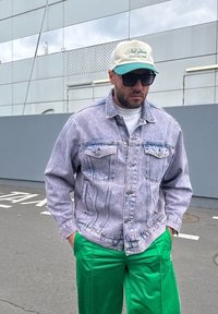 Lilla teksajope kahe esitloka ja valge t-särgiga all, eredalt rohelised püksid ja beež/roheline nokamüts tekstiga. Kandmas päikeseprille.