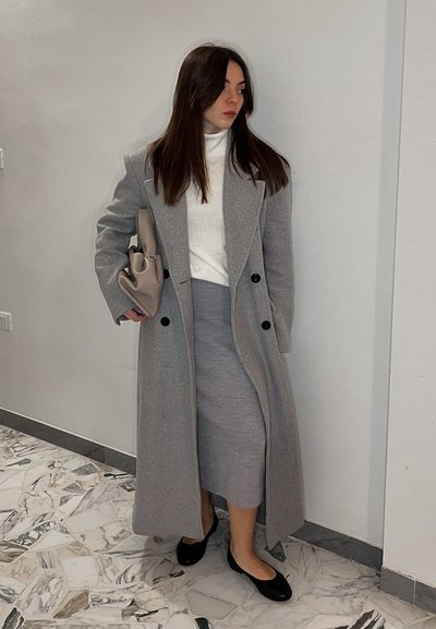 Abrigo largo gris claro con botones negros, suéter de cuello alto blanco, falda lápiz gris y bailarinas negras. Sosteniendo un bolso topo. Fondo de suelo de mármol.