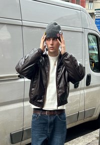 Jeune homme ajustant un bonnet gris avec des patchs, portant une veste en cuir noire, un pull blanc, un jean bleu, debout devant un fourgon blanc dans une rue de la ville.