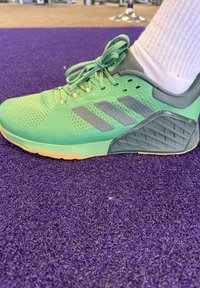 Chaussure de sport verte avec des accents gris, tige en mesh, semelle en caoutchouc, laçage ajustable, avec un talon texturé à motif, sur une surface violette.