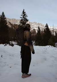 Randonneur avec un grand sac à dos se tient sur un sol enneigé, regardant des montagnes lointaines aux sommets enneigés et des arbres à feuilles persistantes sous un ciel dégagé.