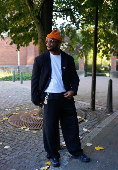 Blazer oversized negro sobre una camiseta blanca, pantalones negros holgados, gorro naranja, gafas redondas y accesorio de cadena; entorno al aire libre con vegetación.