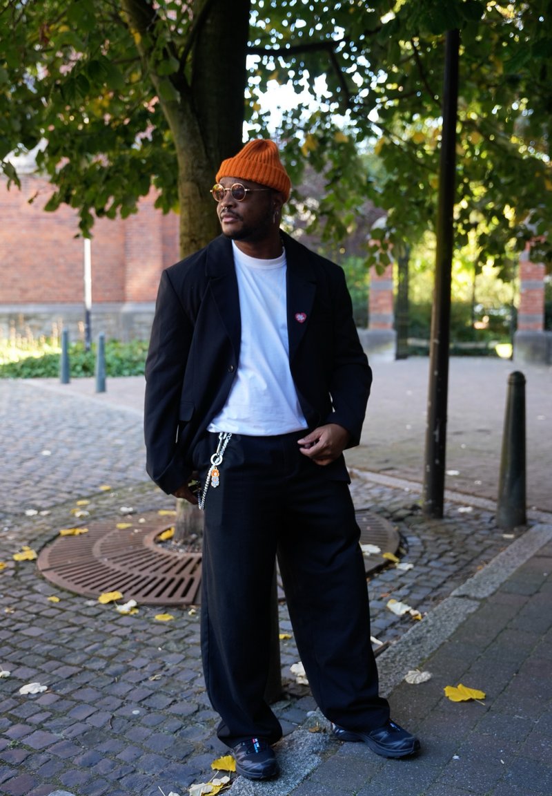 Blazer oversized preto sobre uma t-shirt branca, calças largas pretas, gorro laranja, óculos redondos e acessório de corrente; ambiente ao ar livre com vegetação.