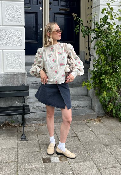 Blusa blanca con estampado floral, falda envolvente de mezclilla oscura, mocasines crema, calcetines tobilleros blancos. La escena presenta escalones al aire libre y vegetación.