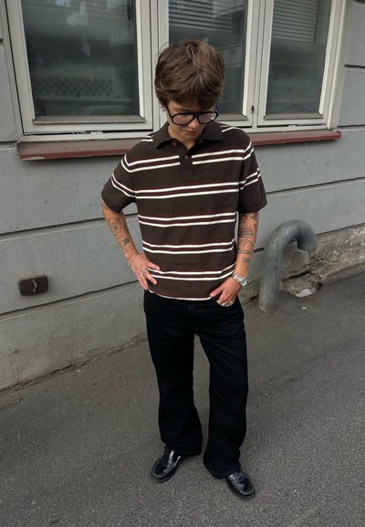 Persona con el pelo corto y gafas, que lleva una camisa marrón con rayas y pantalones negros, de pie con las manos en los bolsillos en una acera.