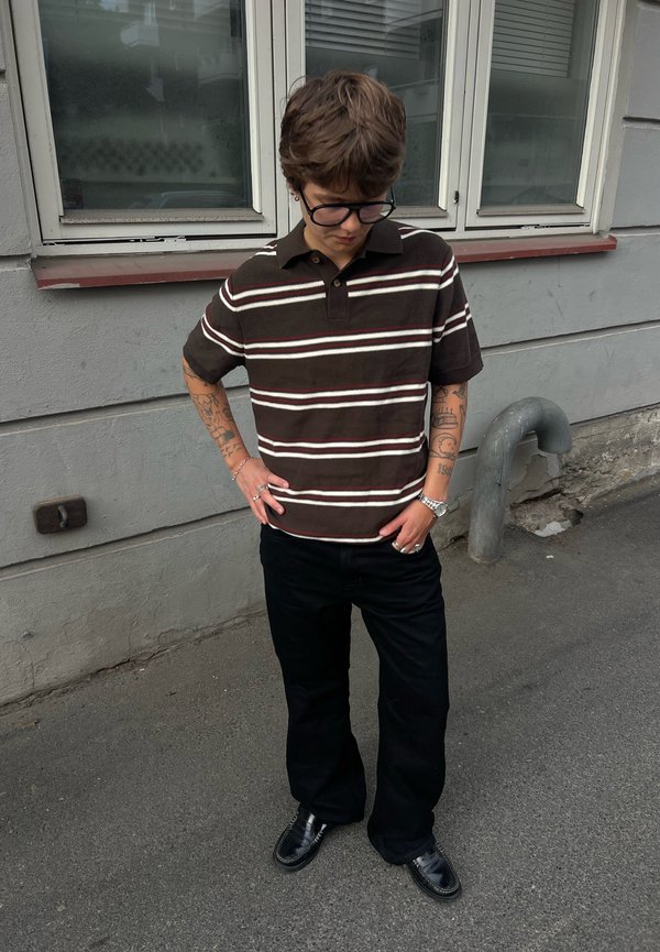 Henkilö, jolla on lyhyet hiukset ja silmälasit, pukeutuneena ruskeaan raidalliseen paitaan ja mustiin housuihin, seisoo kädet taskuissa jalkakäytävällä.