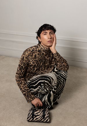 Persona con cabello rizado oscuro en cuclillas, vistiendo una blusa de estampado de leopardo, pantalones de estampado de cebra y sosteniendo un pequeño bolso de estampado de cebra, frente a una pared lisa.