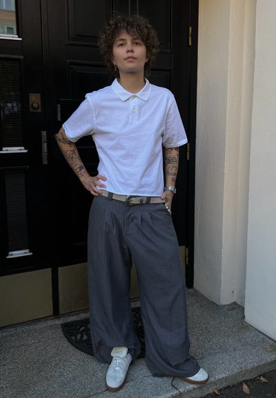 Camisa polo blanca, pantalones de pierna ancha gris con rayas, zapatillas blancas con detalles en marrón, de pie frente a una puerta negra. Tatuajes detallados visibles en los brazos.