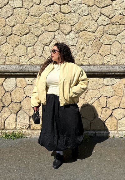 Mujer con cabello rizado que lleva gafas de sol, chaqueta amarillo claro, camiseta crema, falda larga negra, sosteniendo una pequeña bolsa negra, de pie frente a una pared de piedra texturizada.
