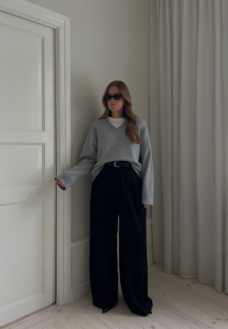 Femme aux longs cheveux portant des lunettes de soleil noires, un pull gris et un pantalon large noir, debout près d'une porte blanche et de rideaux blancs dans une pièce minimaliste.