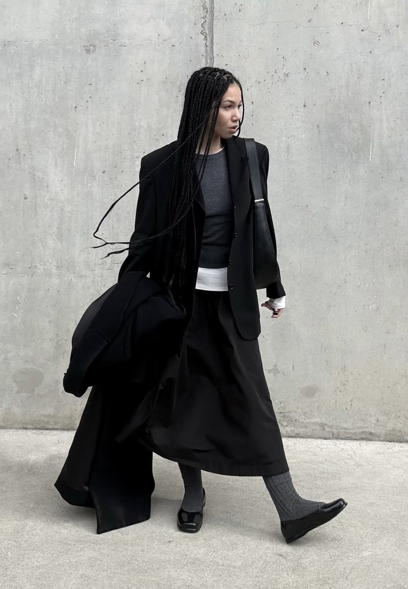 Woman with long braids walking against a concrete wall, wearing layered black and grey clothing and carrying a black jacket and bag.