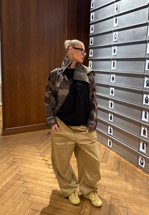 Persona con gafas y chaqueta de camuflaje, camiseta negra y pantalones beige holgados, de pie junto a una pared con fotos en una habitación con suelo de madera.