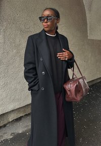 Γυναίκα με μαύρο μακρύ παλτό, μαύρο πουλόβερ, σκούρο παντελόνι, φοράει μεγάλα γυαλιά ηλίου, κρατώντας καφέ δερμάτινη τσάντα, στέκεται μπροστά σε υφή τοίχου.