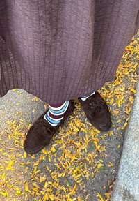 Fødder iført mørkebrune sko og stribede sokker under en struktureret lang nederdel, stående på en jord dækket af gule blade.