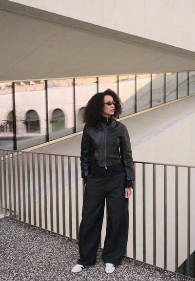 Mujer con cabello rizado, que lleva una chaqueta de cuero negra, pantalones anchos negros, zapatillas blancas y gafas de sol, de pie cerca de una barandilla de un edificio moderno.
