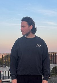 Μαύρη μπλούζα με χαλαρή εφαρμογή και λεπτομέρεια λογότυπου, συνδυασμένη με μαύρο παντελόνι. Το ύφασμα φαίνεται μαλακό και ελαφρώς υφασμένο.