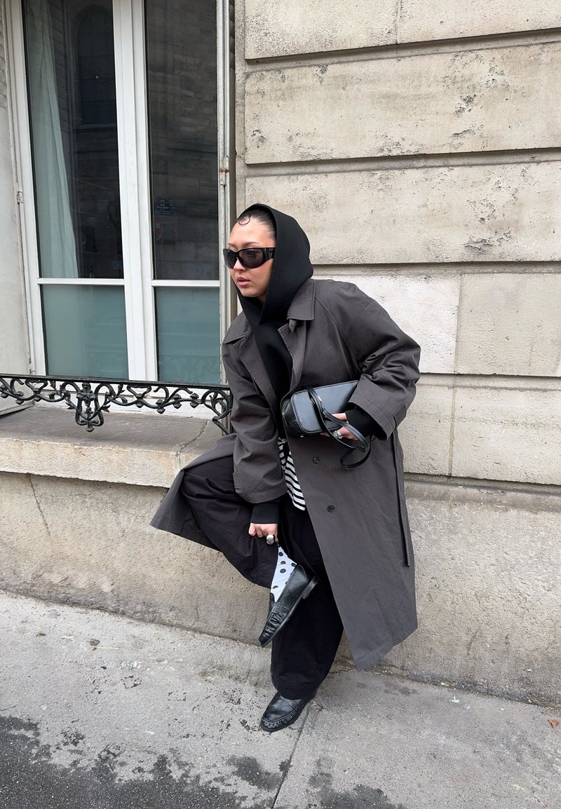 Personne portant un manteau sombre surdimensionné, un sweat à capuche noir, des lunettes de soleil, un pantalon noir, des chaussettes à pois et des mocassins ajuste sa chaussure près d'un mur en pierre.