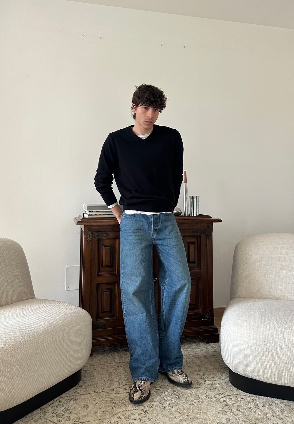 Mand i sort sweater og blå jeans, der læner sig mod et træskab mellem to beige lænestole i en minimalistisk stue.